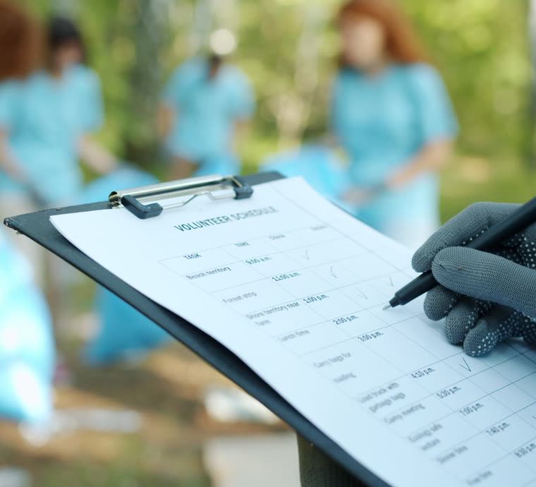 A gloved hand filling in a volunteer schedule on a clipboard, with volunteers blurred behind